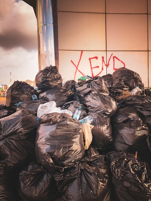 A large pile of black rubbish bags, some slightly torn or bulging, is stacked against an external tiled wall adjacent to a modern building with reflective surfaces. The bags are made of plastic and ap